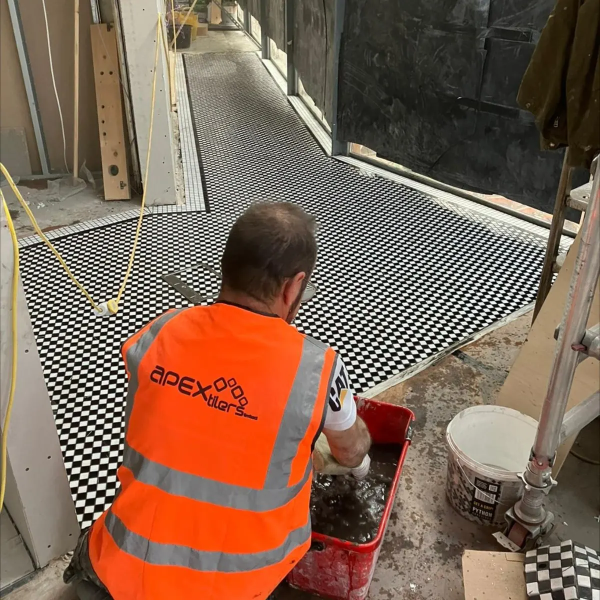 Apex Tilers tiler in branded hi-vis bedding a black and white checkerboard mosaic floor in a London entrance hallway